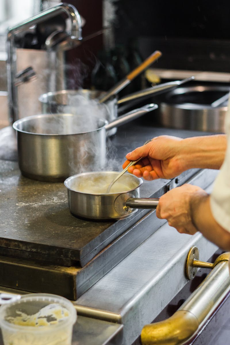 A chef's hands stirring sauce in a stainless steel pan, creating steam in a professional kitchen setting.